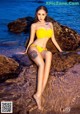 A woman in a yellow bikini sitting on a rock by the water.